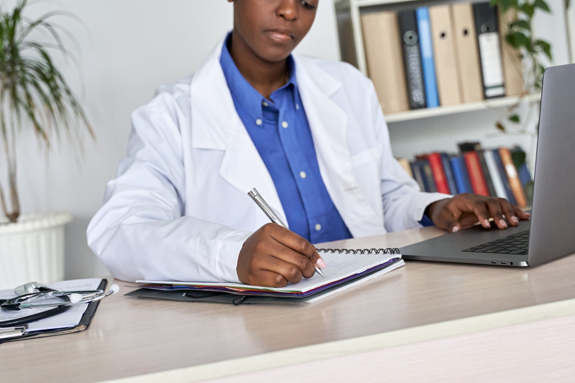 African woman gp doctor wears white coat using laptop writes notes sits at desk.